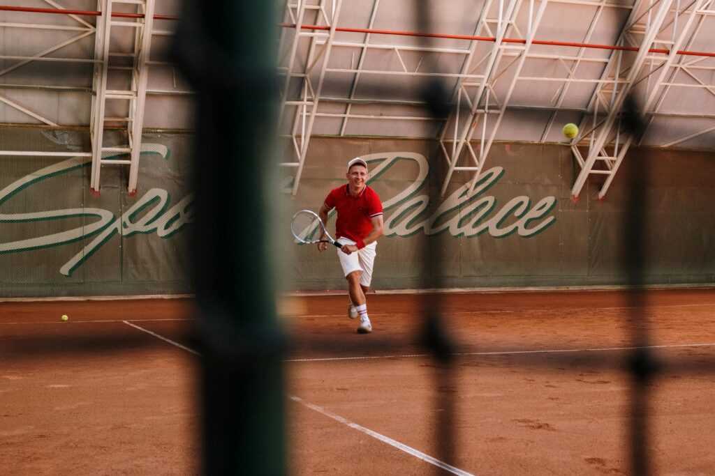 A male tennis player in action on a clay court at Sports Palace indoor facility.